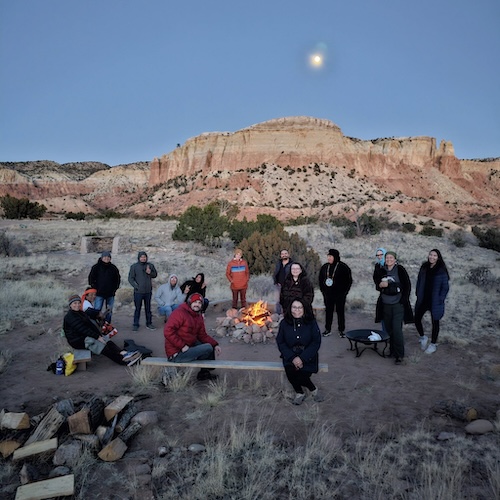 people sitting and standing around a fire with a New Mexican mountain in the background